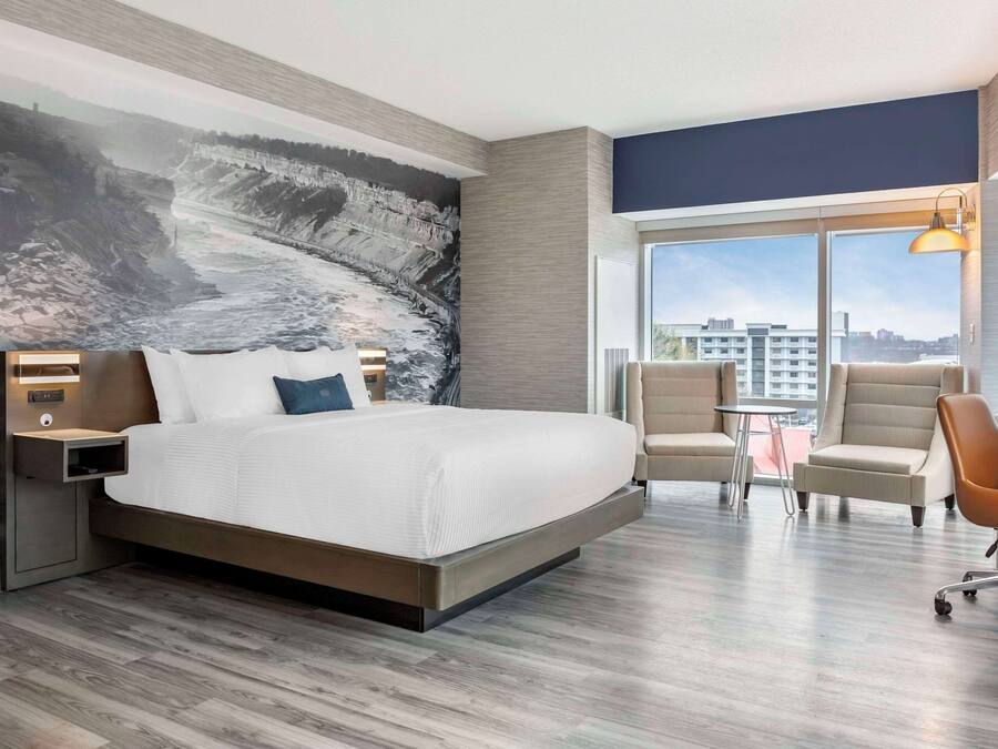 A modern hotel room with a large bed, a desk, two chairs, and a city view through large windows.