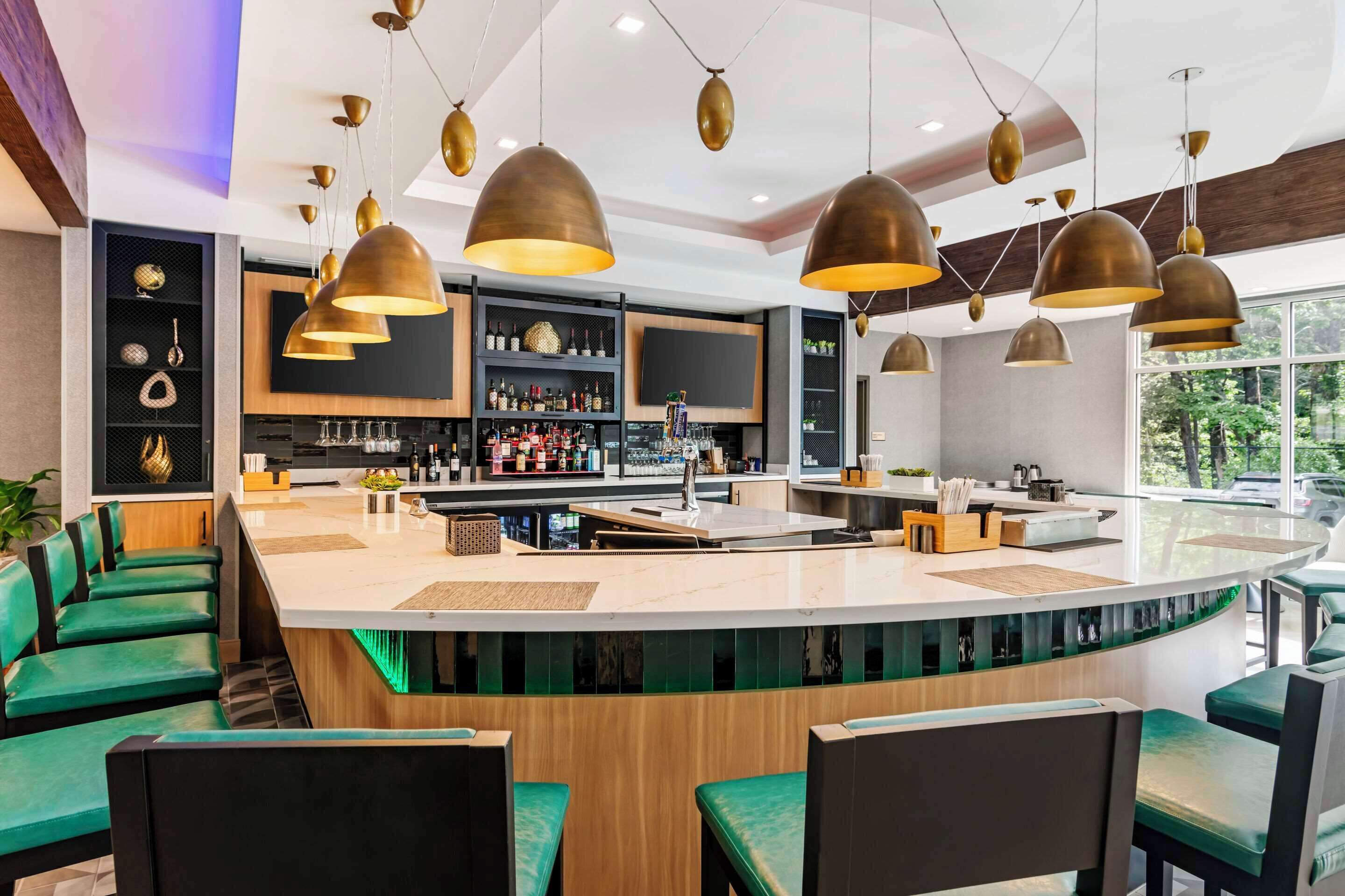 A modern bar area with a curved counter, green upholstered bar stools, and pendant lighting.