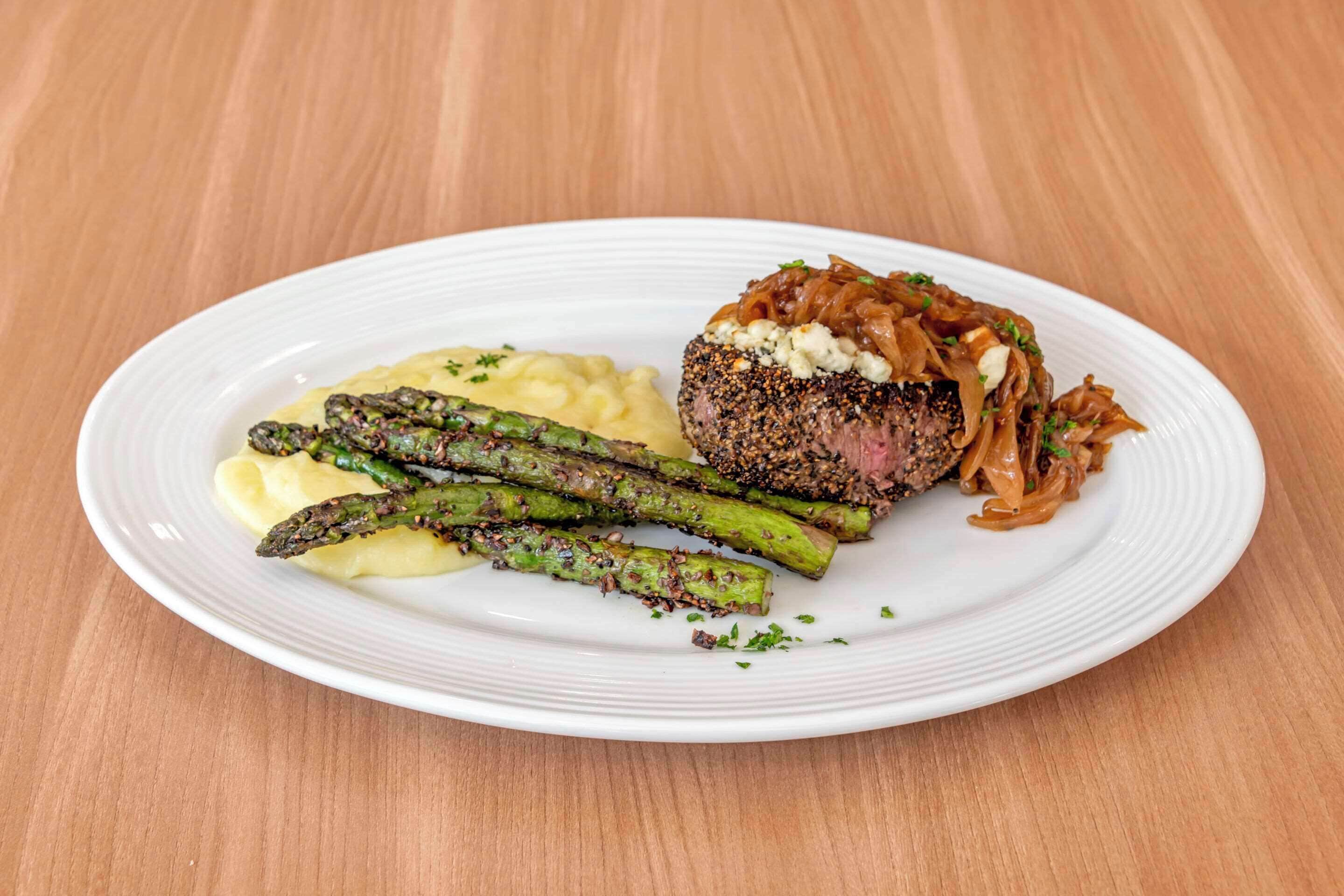 A plate with a steak topped with onions, asparagus, and a creamy sauce, served on a white plate.