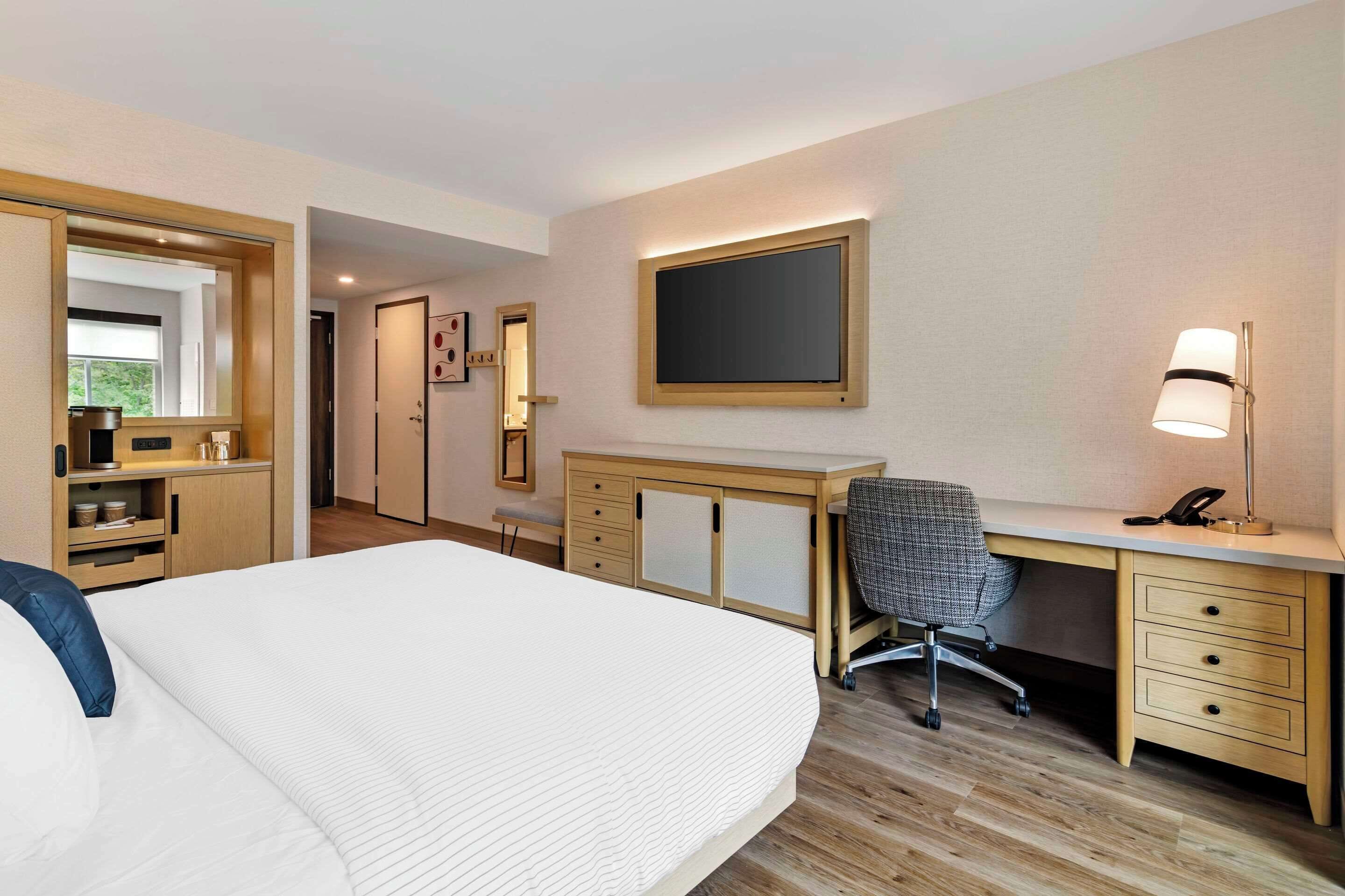 Una habitación de hotel con cama, escritorio, silla, lámpara y armario.