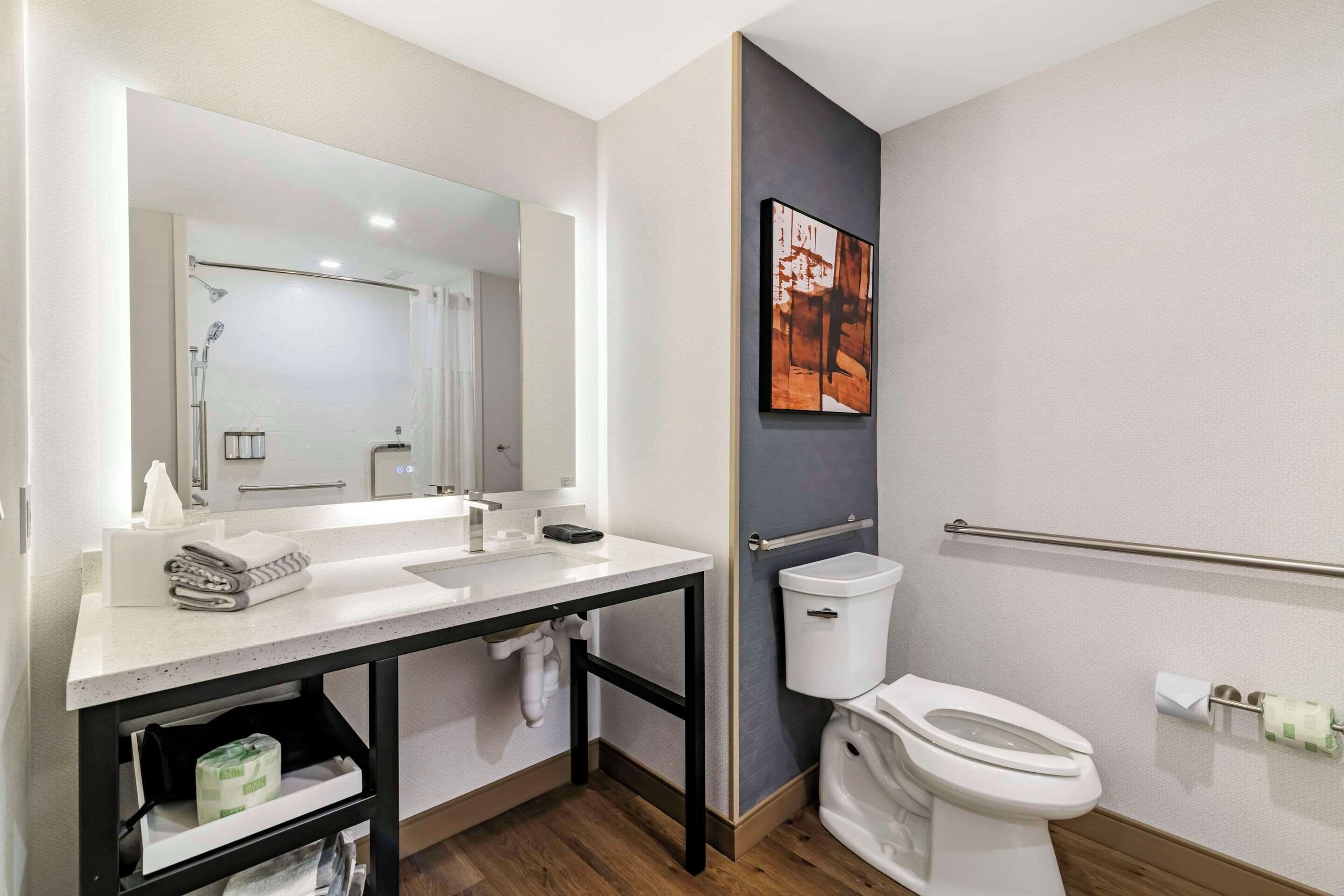 A modern bathroom with a white sink, mirror, and toilet. There is a towel rack and a shelf with toiletries.