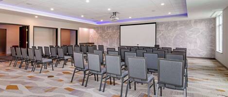 A conference room with rows of chairs, a projector screen, and a patterned carpet.