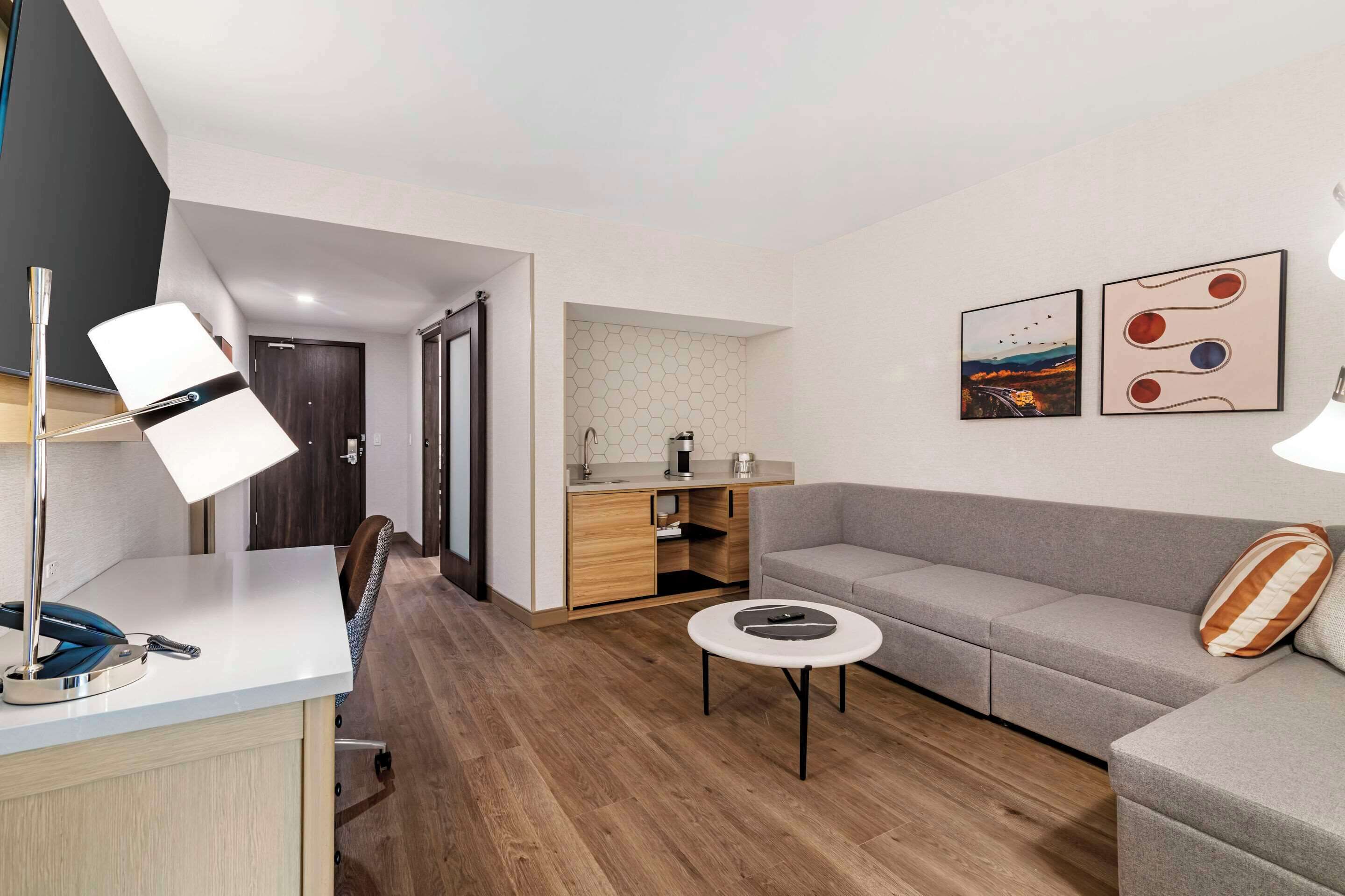 A modern living room with a grey sofa, a small round coffee table, and a desk with a lamp. There is a kitchen area with wooden cabinets and a microwave. Two artworks are displayed on the wall.