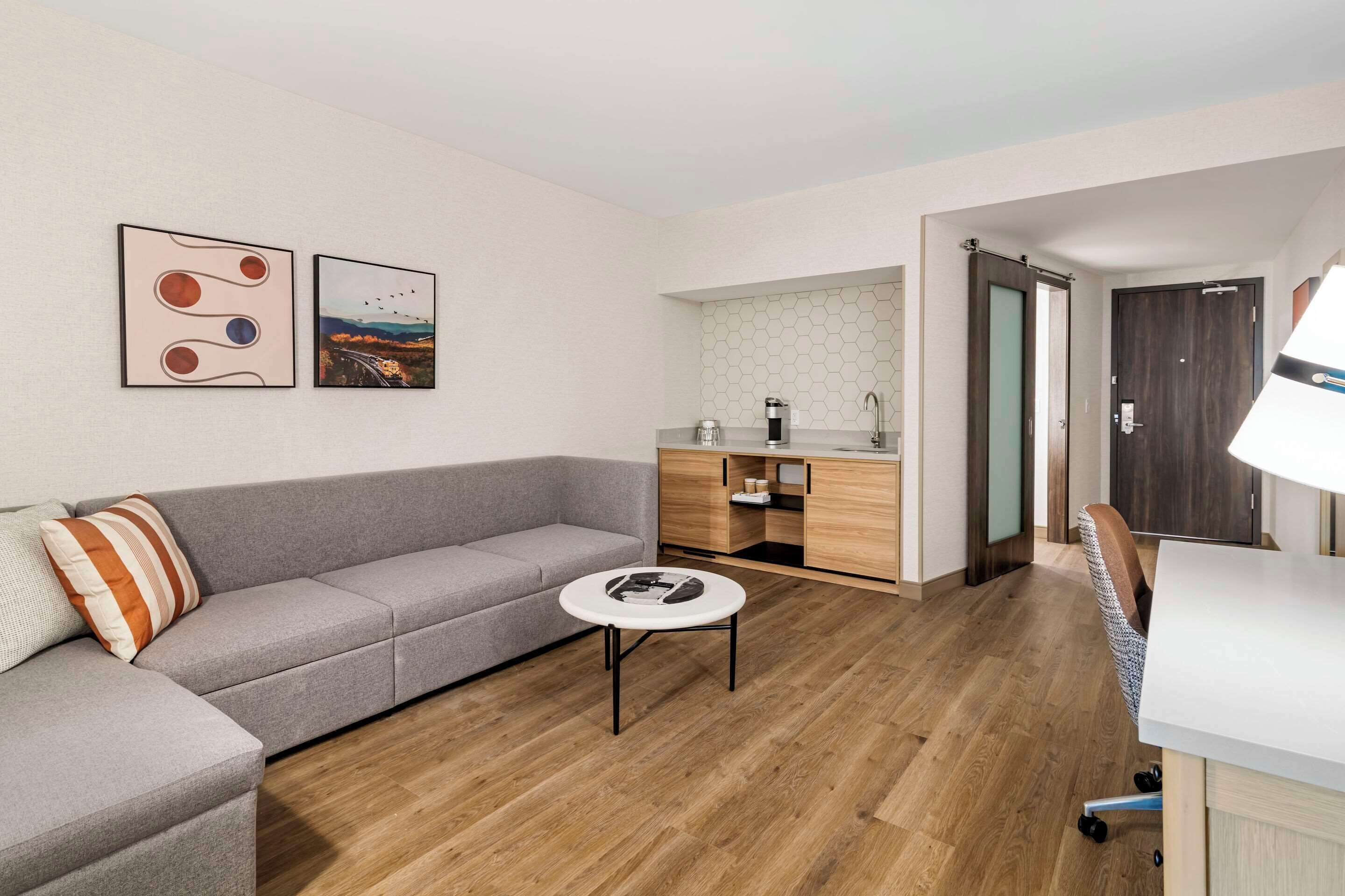 Uma sala de estar moderna com um sofá cinzento, uma mesinha redonda e uma área de cozinha com armários de madeira e bancada branca.