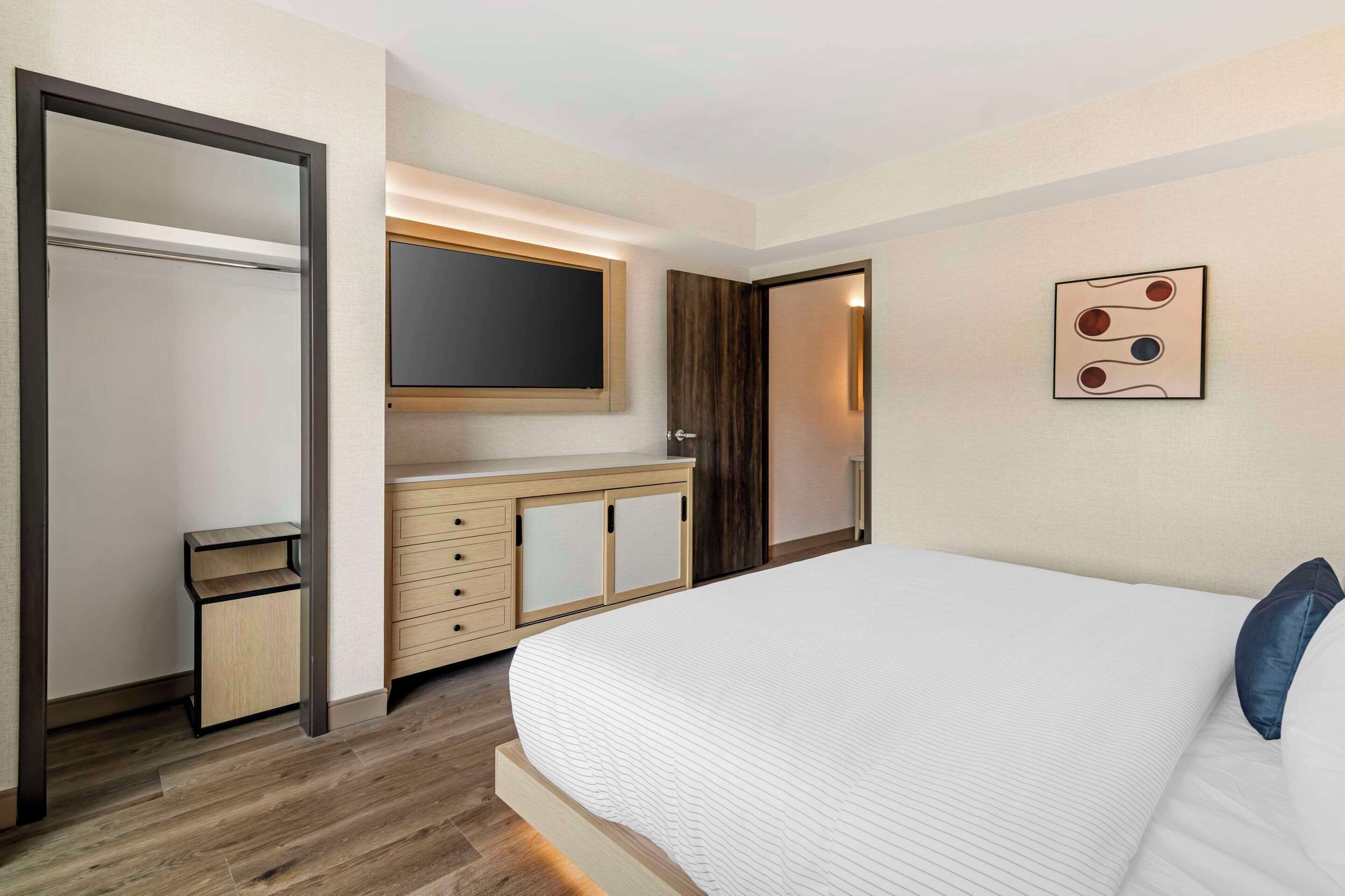A hotel room with a large bed, a wardrobe, a mirror, a flat-screen TV, and a wall-mounted artwork.
