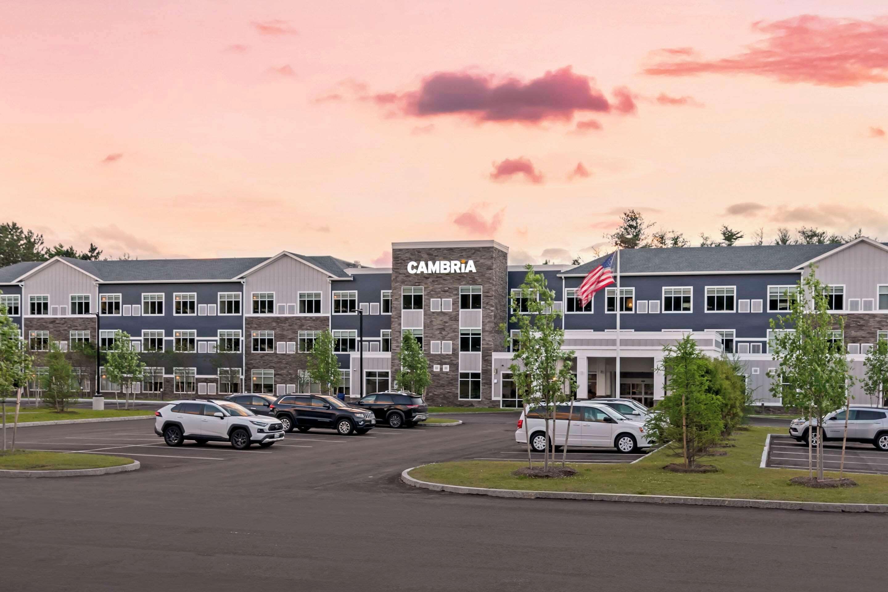 A multi-story building with a sign reading "CAMBRIA" and an American flag. There are several cars parked in front, and the sky is pinkish at dusk.