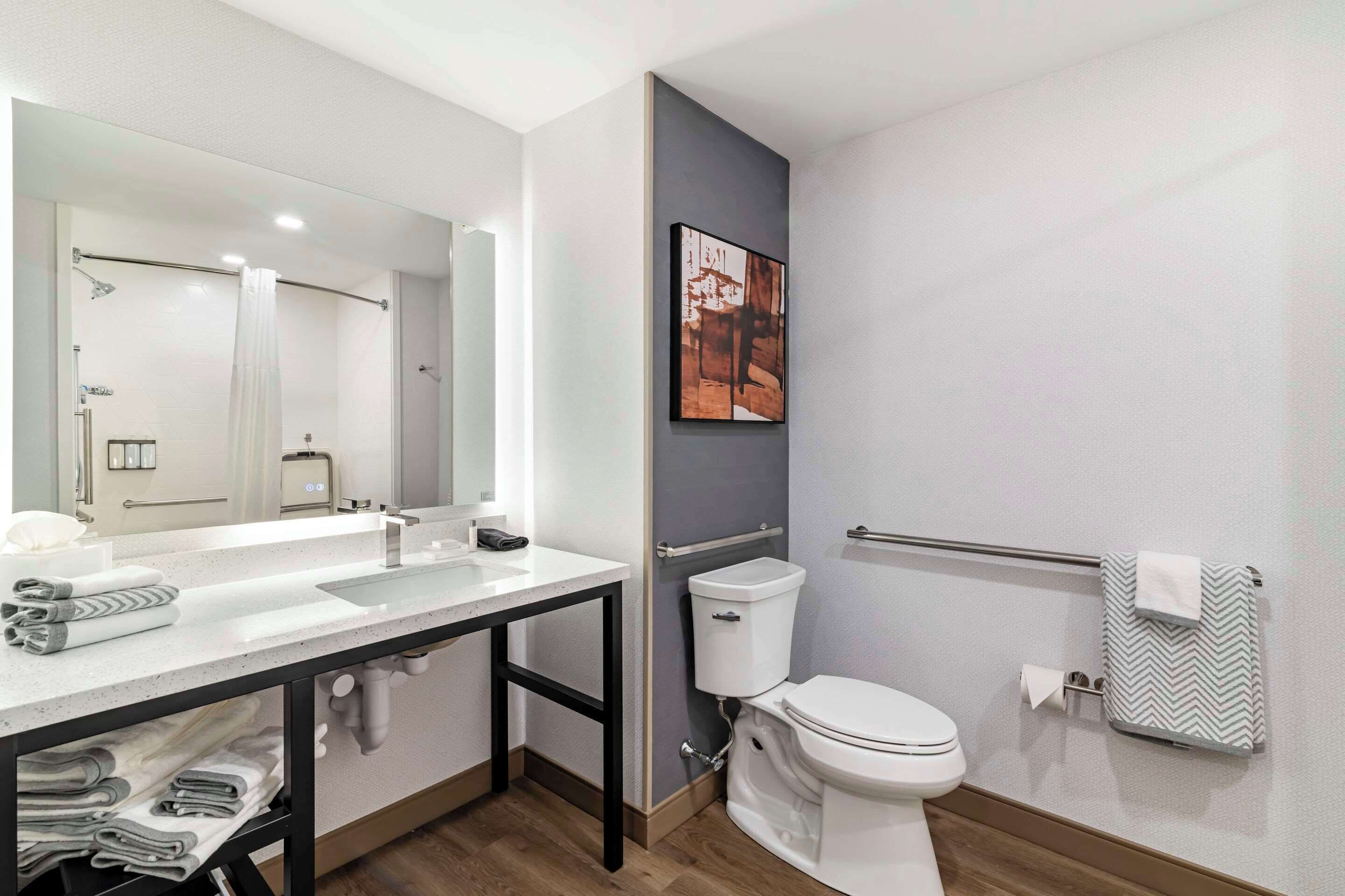 Un baño moderno con un espejo grande, lavamanos blanco y retrete. Hay un perchero con una toalla y un cuadro en la pared.