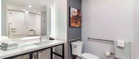 Un baño moderno con un espejo grande, lavamanos blanco y retrete. Hay un perchero con una toalla y un cuadro en la pared.