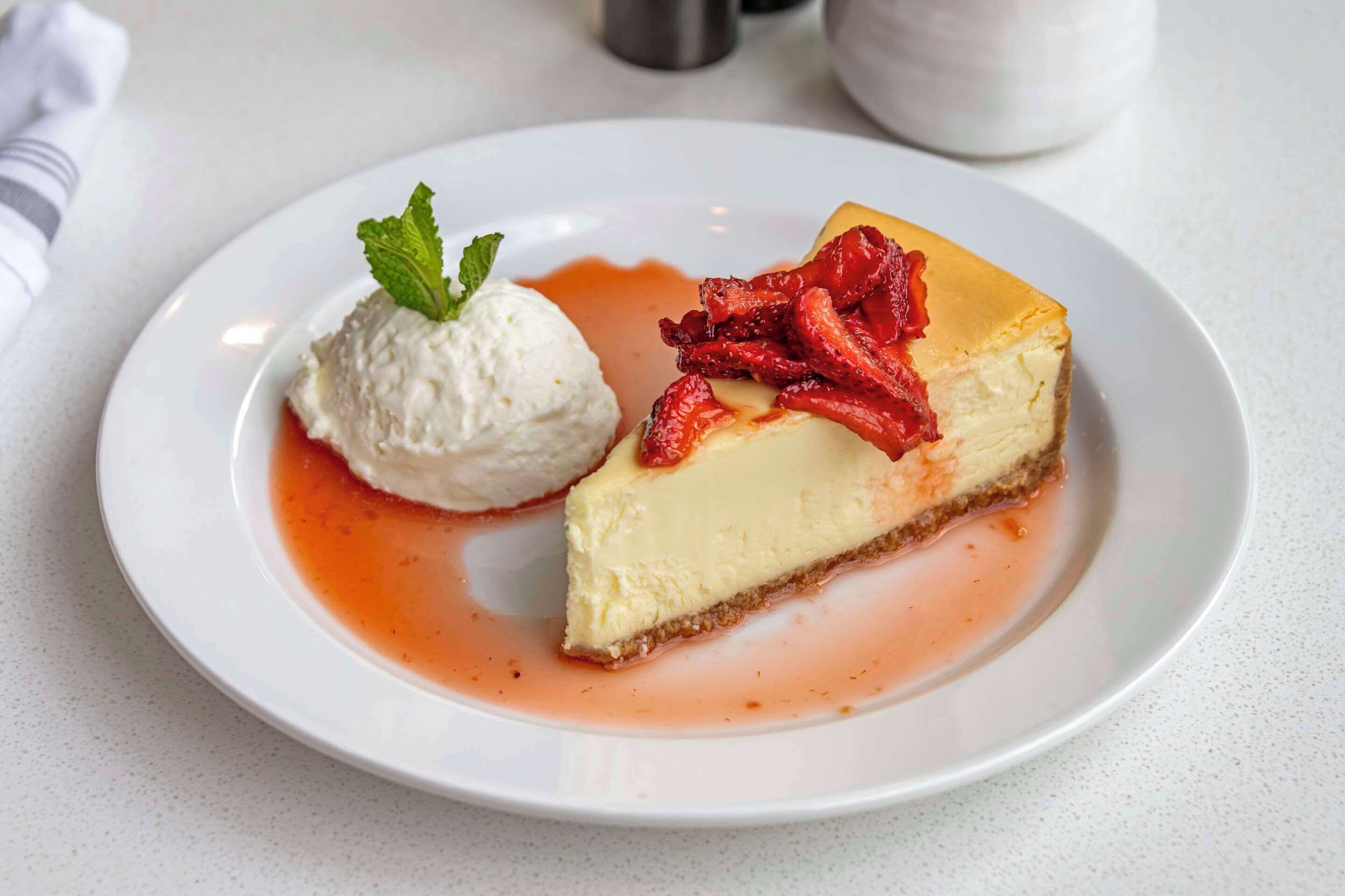 Ein Stück Käsekuchen mit Erdbeeren und eine Kugel Vanilleeis auf einem Teller.