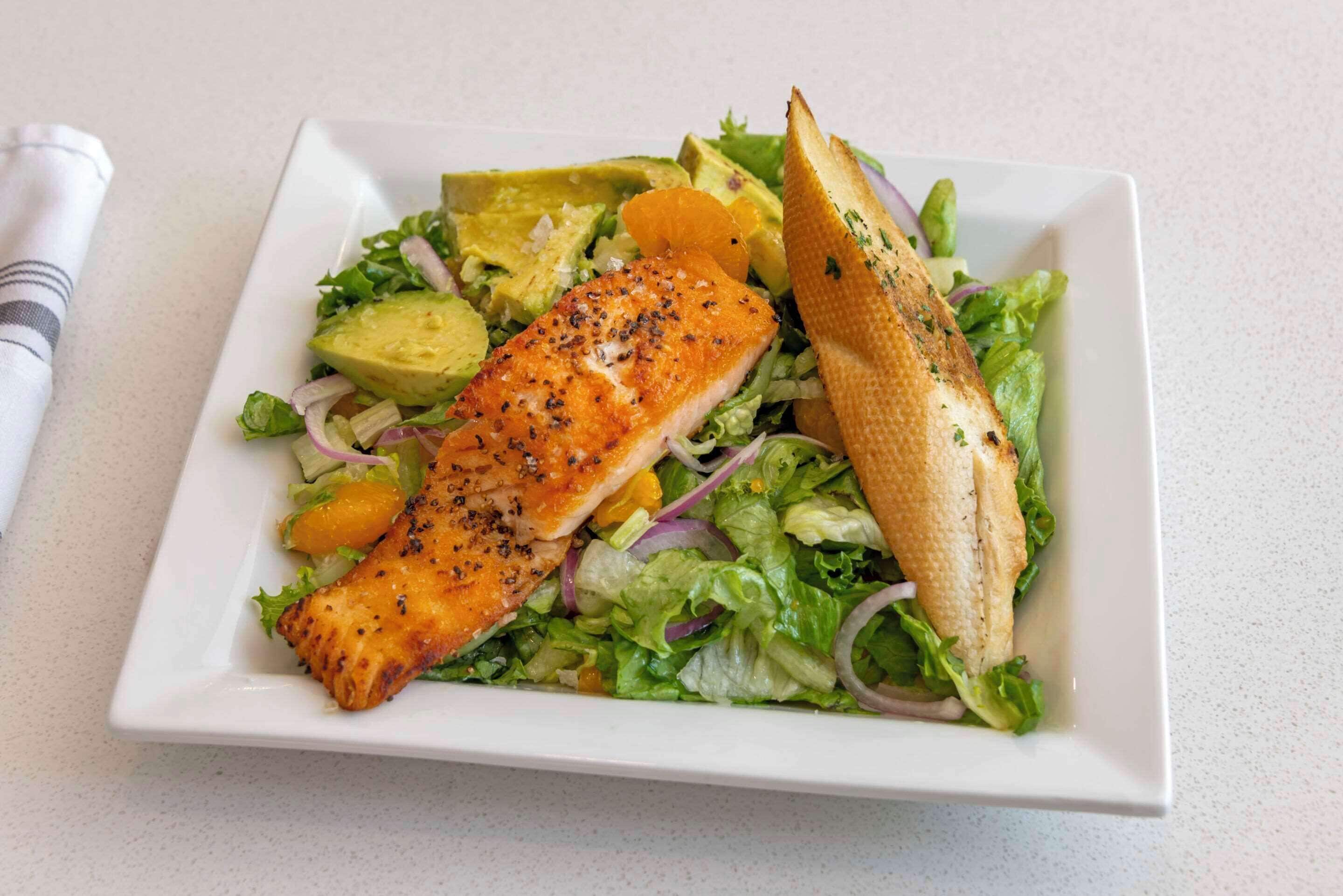 A white square plate with a grilled salmon fillet, avocado slices, and a toasted bread piece on a bed of mixed greens and red onions.