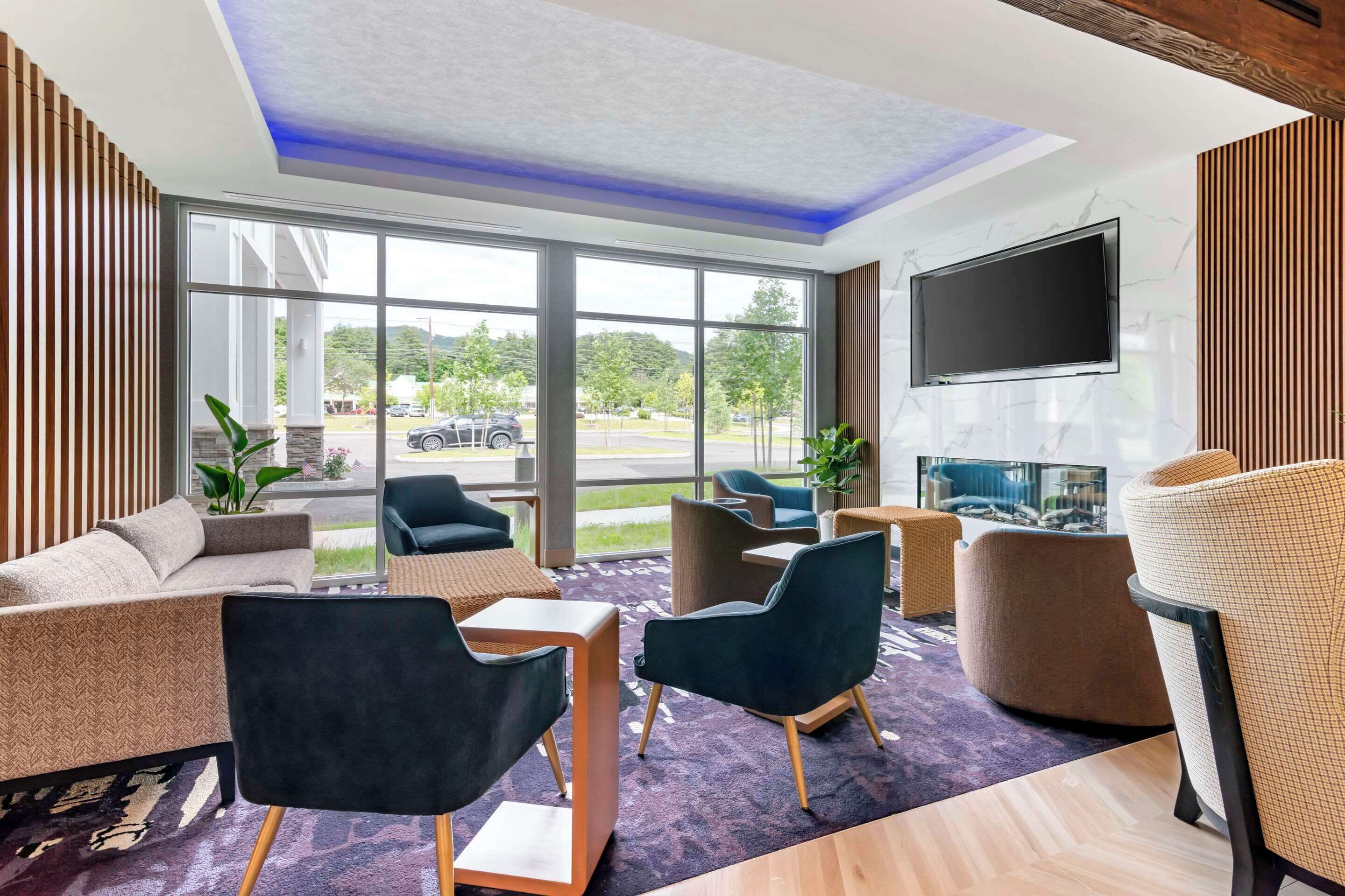 Un moderno vestíbulo de hotel con un sofá, sillas, un televisor de pantalla plana y grandes ventanales con vistas al exterior.