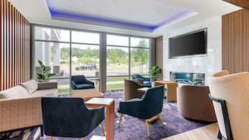 Un moderno vestíbulo de hotel con un sofá, sillas, un televisor de pantalla plana y grandes ventanales con vistas al exterior.