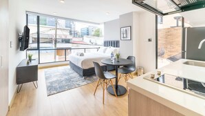 A modern hotel room with a bed, a dining area with a table and chairs, a kitchen area, and a large window offering a city view.