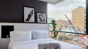 A hotel room with a large bed, white bedding, two framed pictures on the wall, a potted plant, and a view of the cityscape through the window.