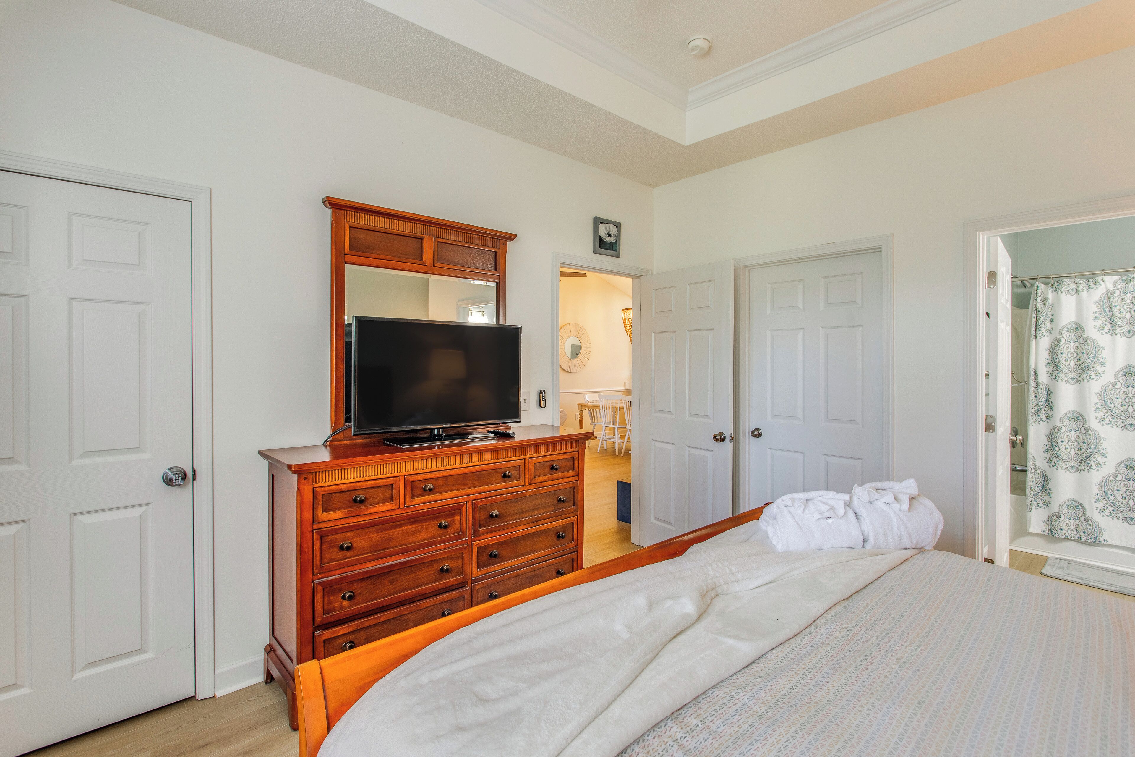 A bedroom with a wooden dresser, a television, a bed with white bedding, and a door leading to a bathroom with patterned curtains.