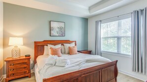 A bedroom with a wooden bed, a nightstand with a lamp, a window with blinds, and a wall painting. - Resort Condo w/ Pool Access Near Barefoot Landing! (North Myrtle Beach)