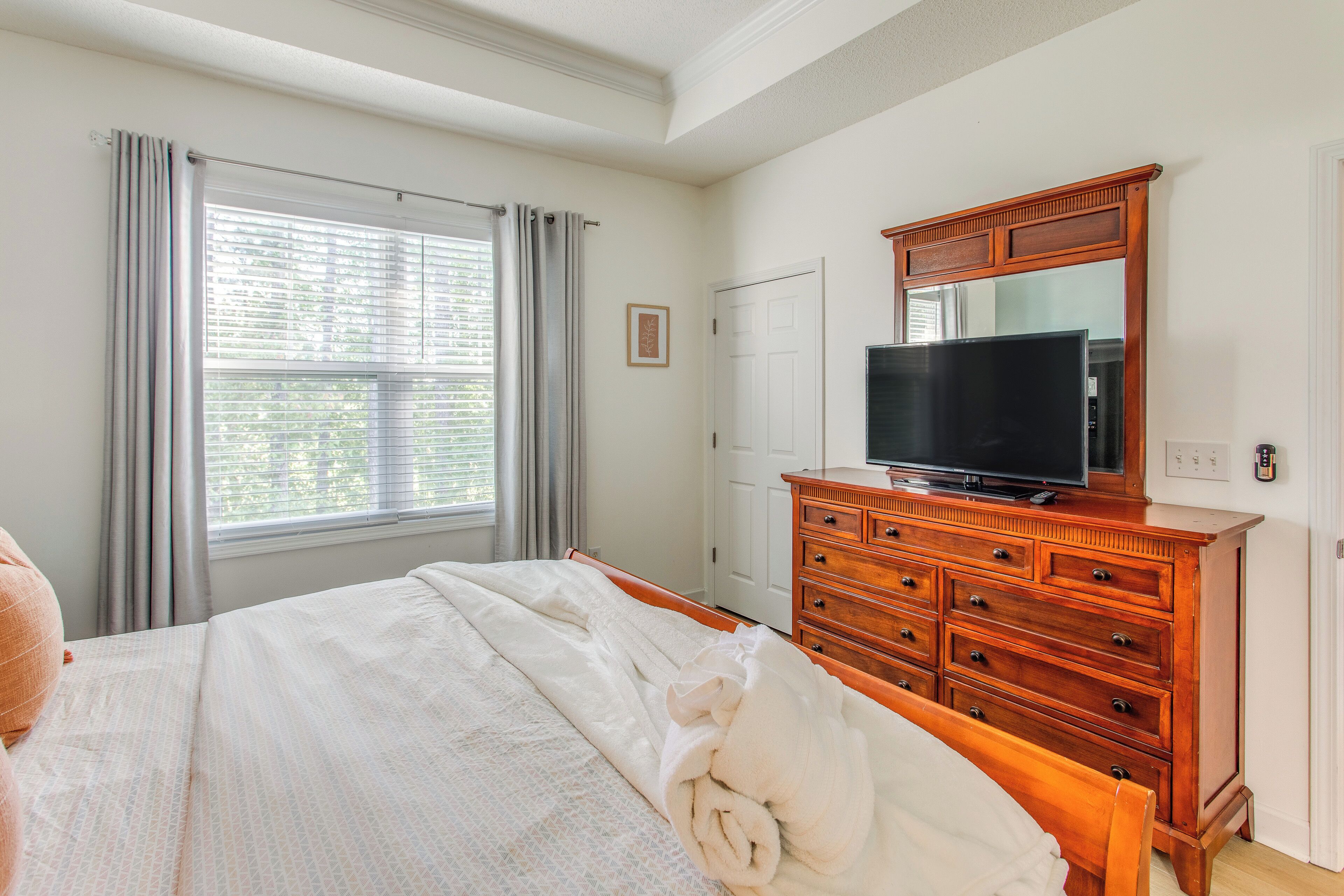 Une chambre à coucher avec un lit, une commode en bois sur laquelle est posée une télévision, une fenêtre avec des rideaux et une porte.