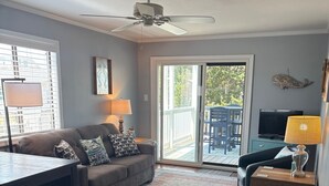 Ein Wohnzimmer mit einer Couch, einem Couchtisch, einem Fernseher und einem Deckenventilator. Eine Glastür führt zu einer Terrassenanlage im Freien.