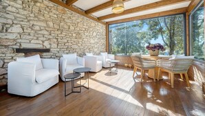 A room with stone walls, wooden beams, white sofas, a dining table with chairs, and a large window with a view of trees. - Entire holiday home 'Mas De L'Arlequi' with Mountain View, Private Pool and Wi-Fi (Rojals)