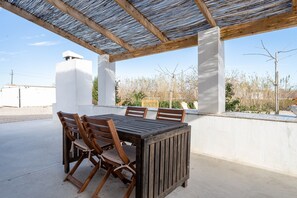 An outdoor patio with a wooden table and chairs, a pergola overhead, and a view of the surrounding landscape.