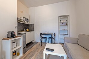 A compact living space with a kitchenette, a dining area with two chairs, a white sofa, and a small table with a remote control. - Apartment 'Les Capucins - T2 Tout Confort' with Private Terrace, Private Garden and Wi-Fi (Brest)