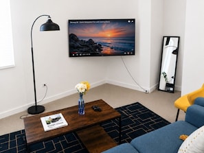 A living room with a TV displaying a scenic film, a coffee table with a remote and a guidebook, a floor lamp, a sofa, and a mirror.
