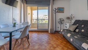 Un salon comprenant un canapé, une table à manger avec des chaises, un téléviseur fixé au mur, un ventilateur et une vue sur l’extérieur, accessible par une porte-fenêtre coulissante.