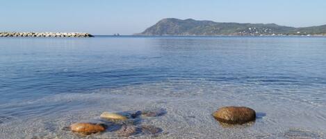 Mar calmo com águas cristalinas, rochas na costa e montanhas ao longe.