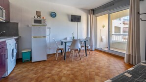 Une cuisine compacte équipée d’un lave-linge, d’un four à micro-ondes et d’un petit réfrigérateur. On y trouve également un coin repas avec une table et des chaises, ainsi qu’une grande fenêtre donnant sur l’extérieur.