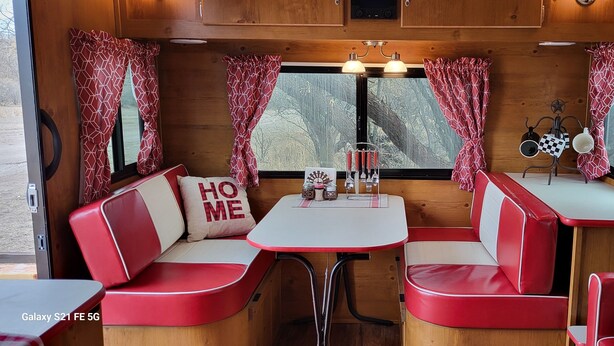 Interior acogedor de una caravana con asientos rojos y blancos, una pequeña mesa y una ventana con cortinas.