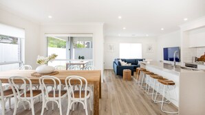 A modern kitchen with a built-in oven, a dining area with a wooden table and white chairs, and a living area with a blue sofa and a TV. - The Coast Beach - Pet Friendly - 2 Mins Walk to Bay (Callala Bay)