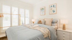 A bedroom with a bed, bedside tables, a window with blinds, and two wall-mounted pictures. - The Coast Beach - Pet Friendly - 2 Mins Walk to Bay (Callala Bay)