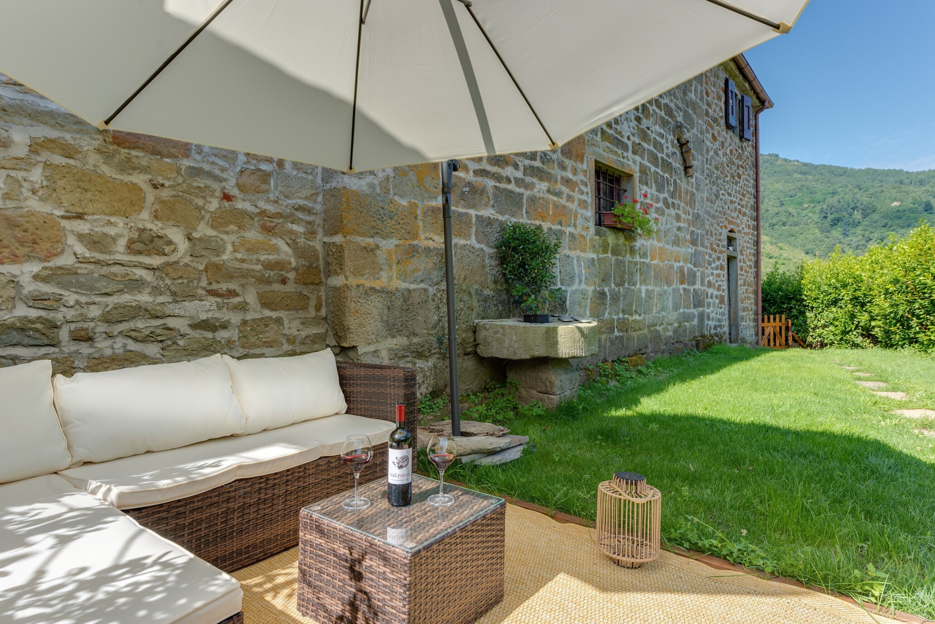 A stone building with a stone wall, a wicker sofa, a table with wine glasses, and a grassy yard.