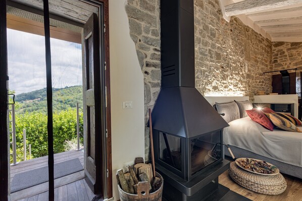Ein Zimmer mit Steinwand, Kamin, Bett mit Kissen und Blick ins Grüne.