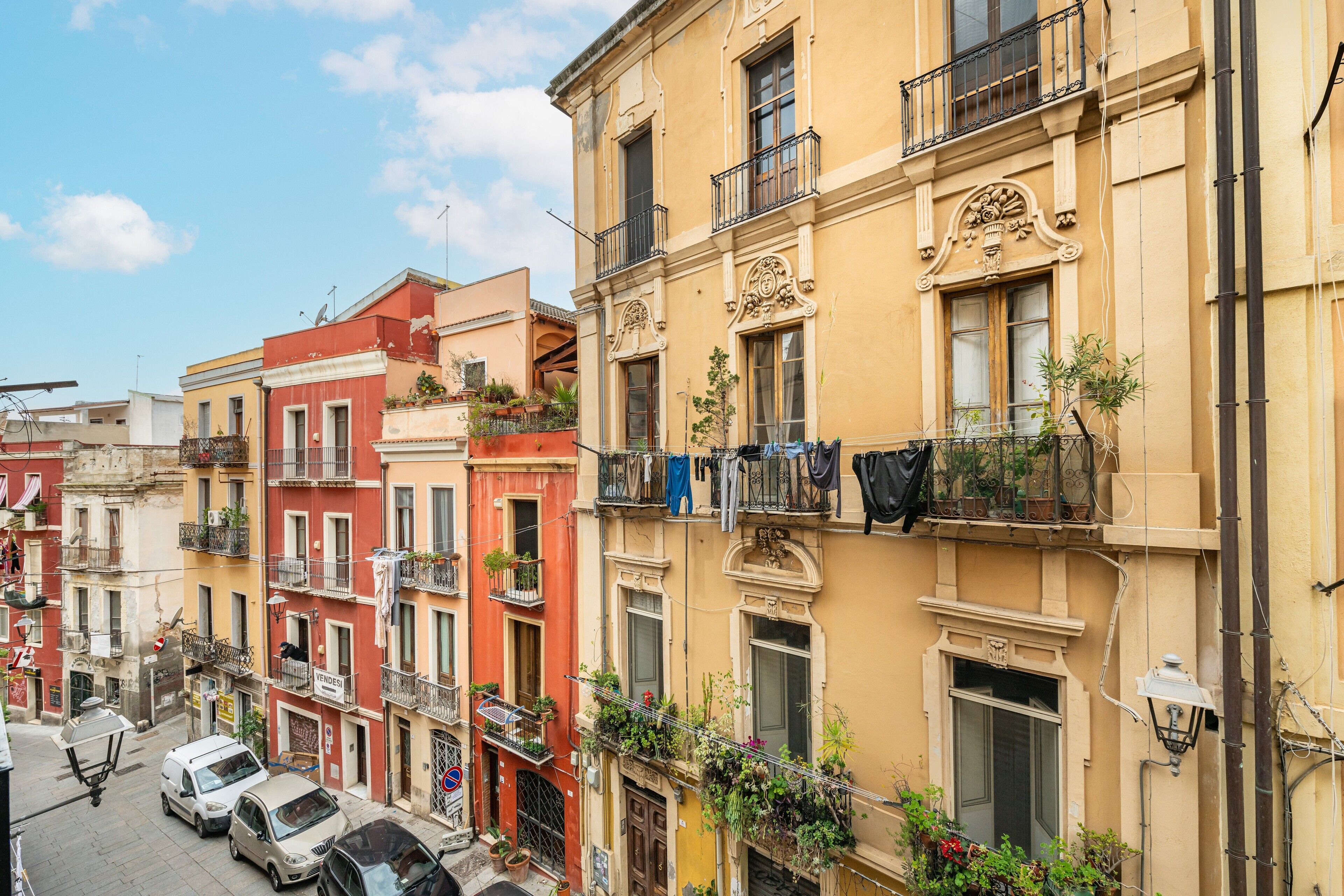 Une rangée d’immeubles à étages avec des balcons, dont certains sont ornés de plantes, et une rue bordée de voitures garées.
