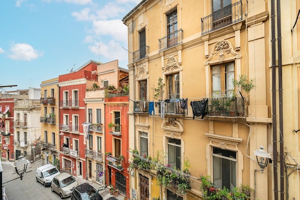 A row of multi-story buildings with balconies, some with plants, and a street with parked cars.
