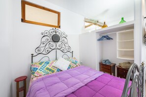 Un lit avec une tête de lit en métal, une courtepointe violette et des coussins colorés. On y trouve une petite table de chevet en bois avec une lampe, un tabouret rouge et une étagère avec des serviettes pliées.