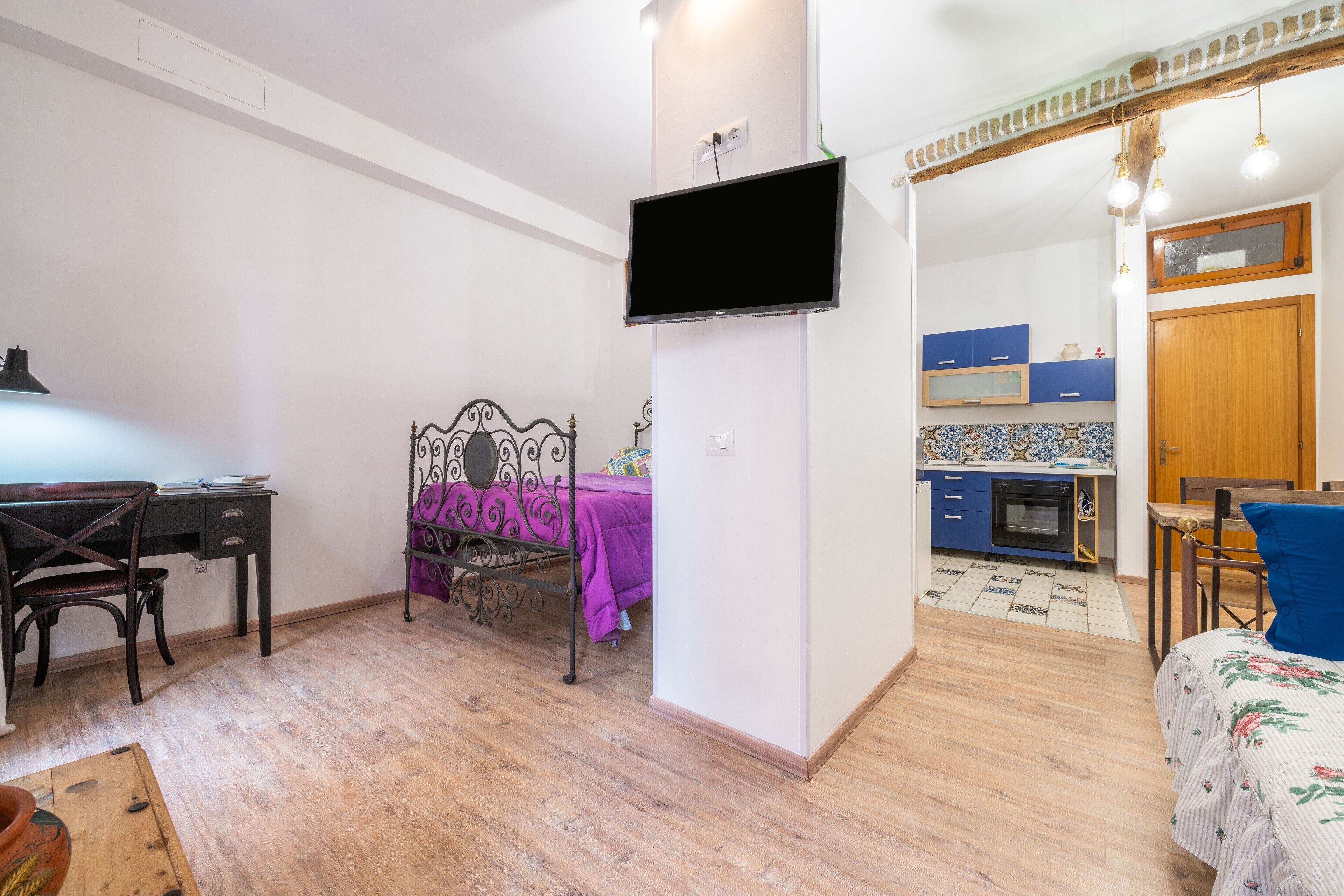A bedroom with a bed, a desk, a chair, a TV, and a kitchen area.