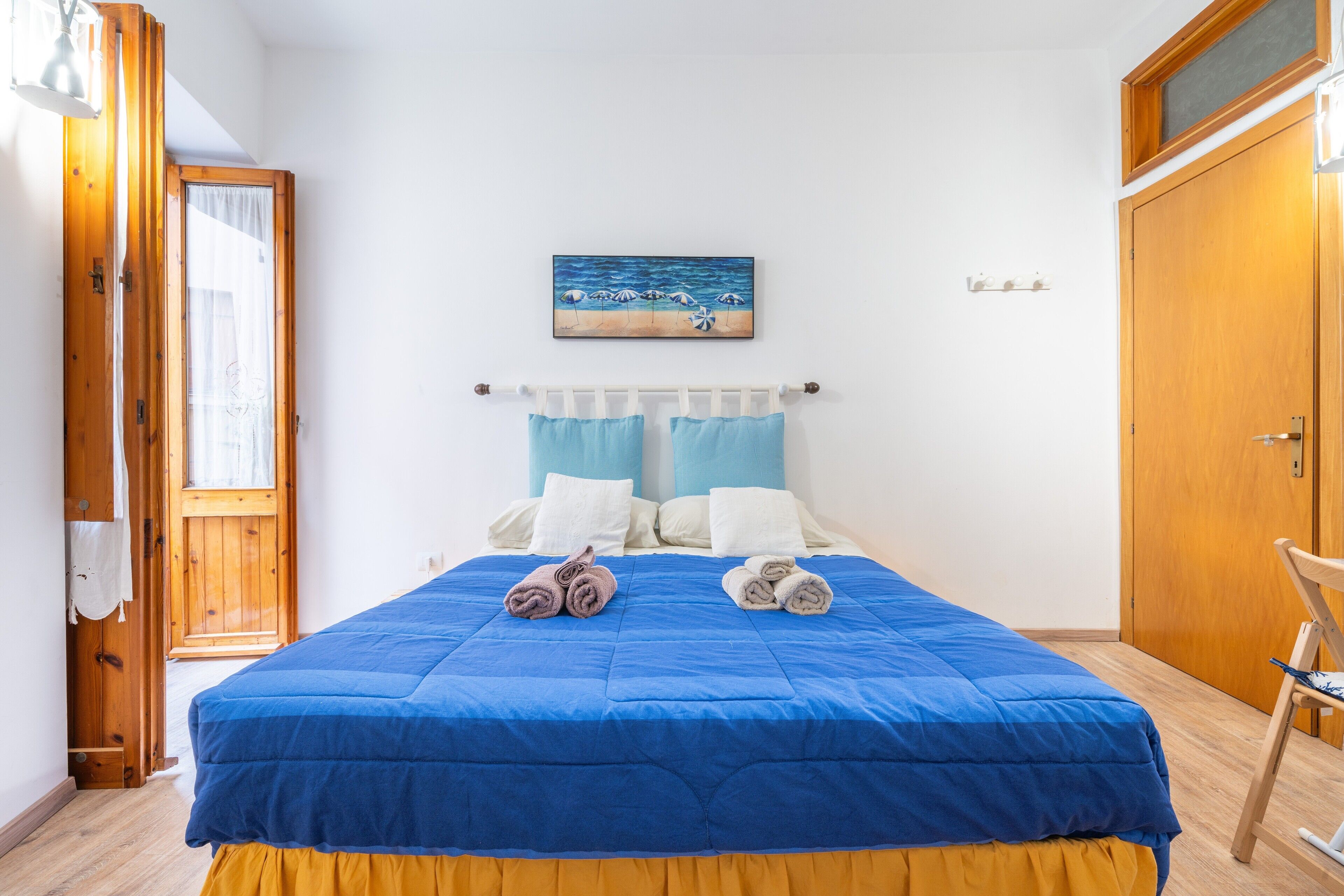 Una camera da letto con una coperta blu, un quadro che raffigura una spiaggia, una porta in legno e una sedia.