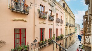 Une rue étroite bordée d’immeubles dotés de balcons ornés de plantes, de fenêtres à encadrement rouge et surplombant un ciel dégagé.