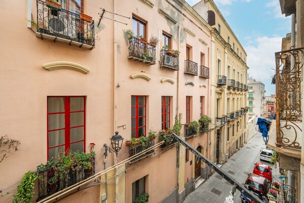 Una calle estrecha con edificios que tienen balcones con plantas, ventanas con marcos rojos y un cielo despejado.