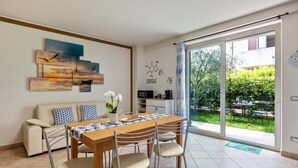 A dining area with a wooden table and chairs, a sofa, and a view of greenery through a glass door. - Bright  apartment 'Bluemoon' with Pool, Wi-Fi and Air Conditioning (LAZISE)