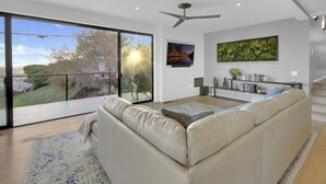 A modern living room with a large sliding glass door leading to a balcony, a ceiling fan, and a bookshelf with books and decorative items. - The Gem of Mission Hills: Luxury 5BR Home with Luxe Amenities and views` (San Diego)