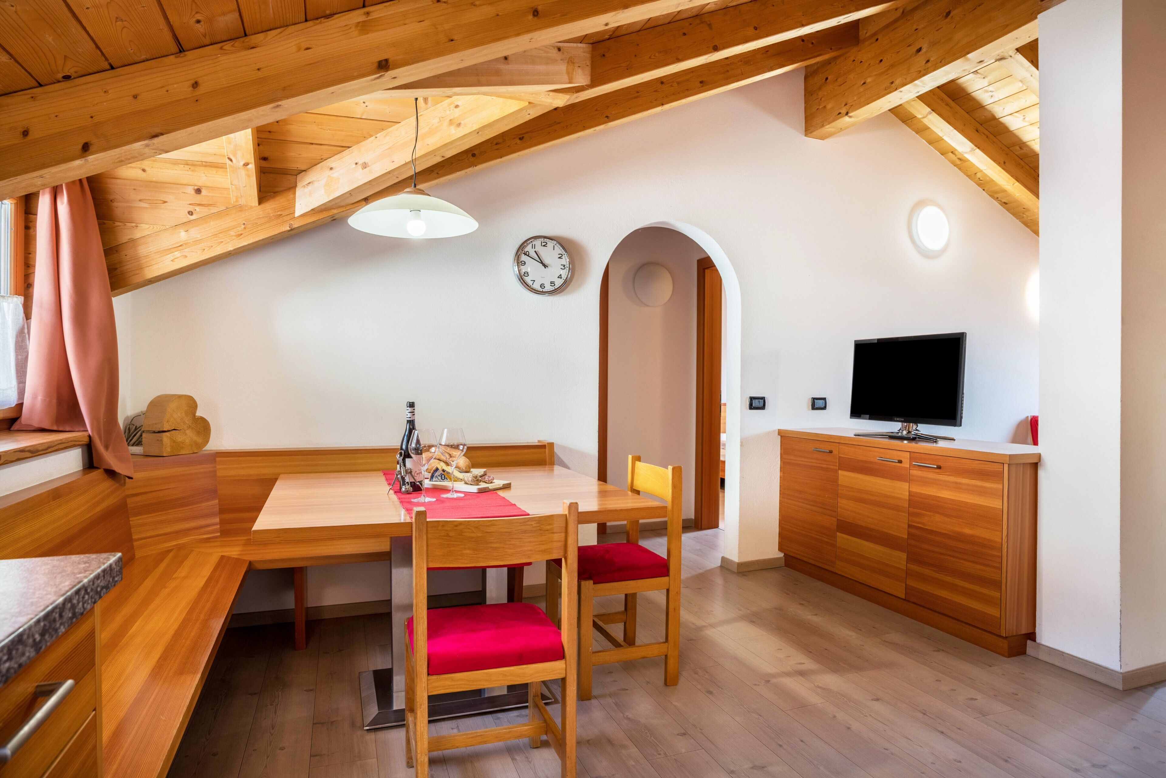 A dining area with a wooden table, chairs, a clock, and a television on a wooden cabinet.