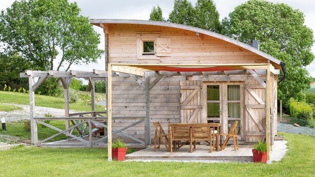 A wooden cabin with a covered porch, a table and chairs, and two potted plants.