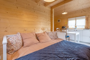 A wooden cabin interior with a bed, a white desk, and a window.