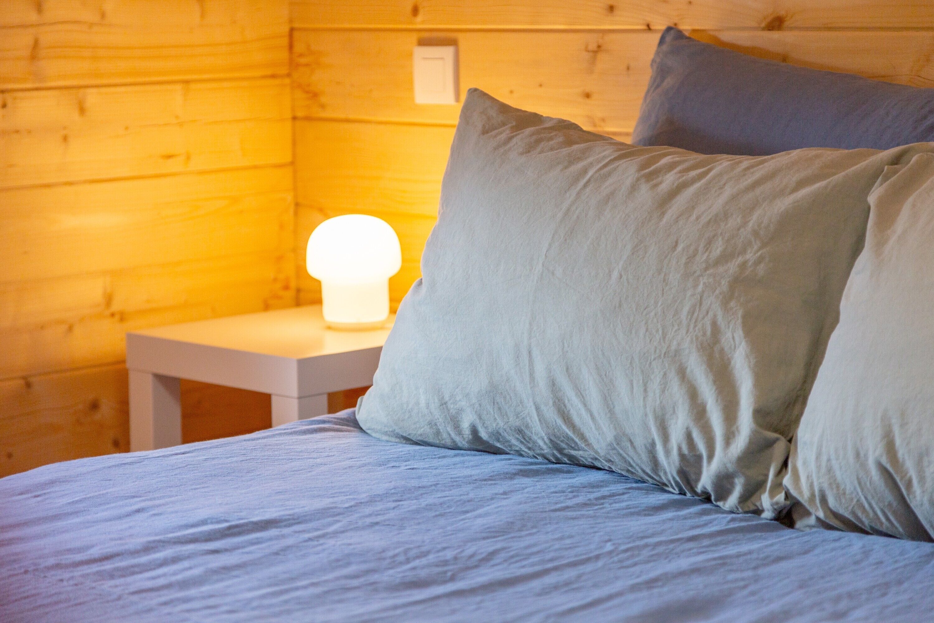 Interior de una cabaña de madera con cama, almohadas y mesita de noche con lámpara.