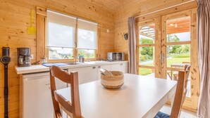 A wooden cabin interior with a kitchen, dining table, and a view of the outdoors.