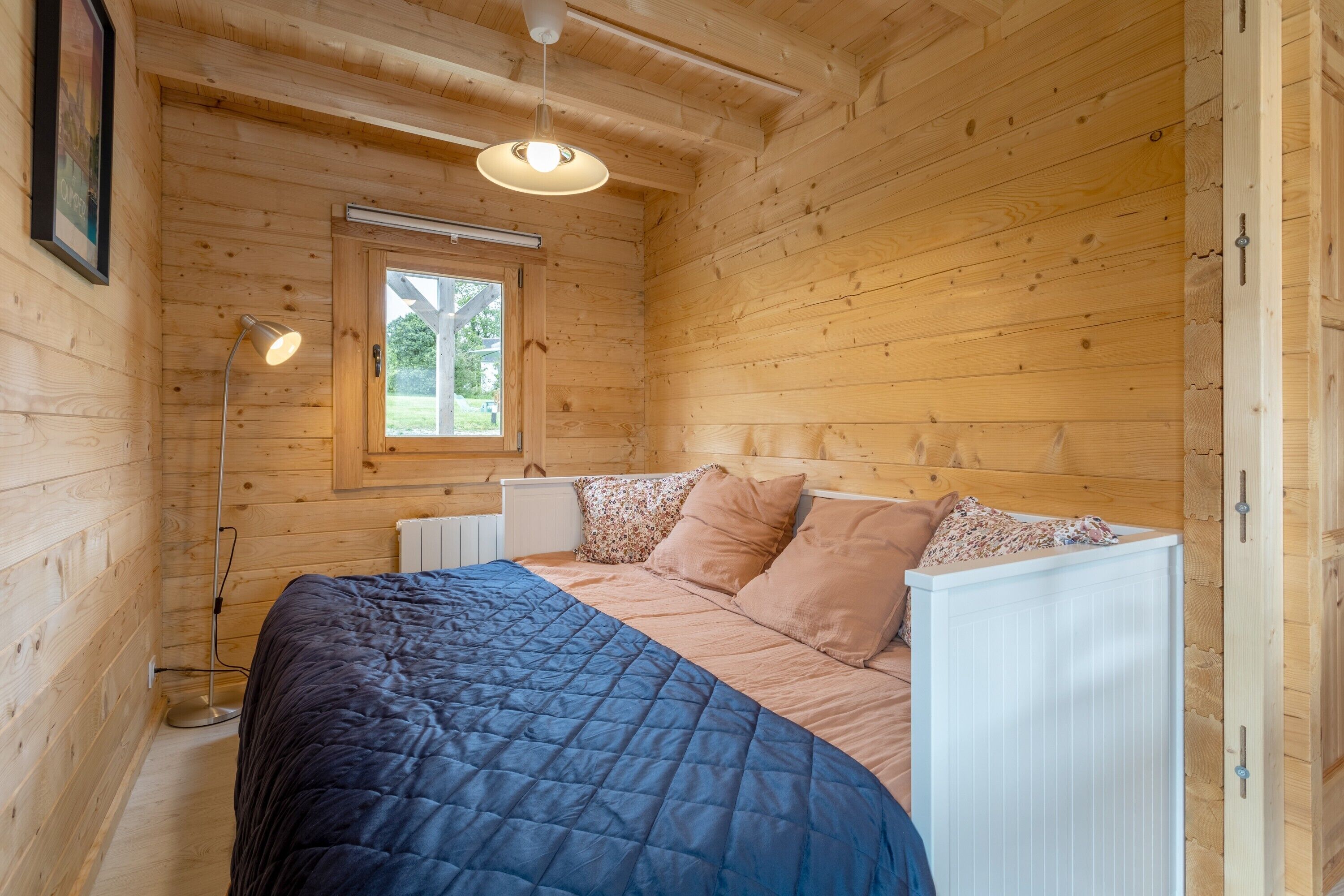Un dormitorio tipo cabaña de madera con una cama, una ventana, una lámpara de pie y un radiador.