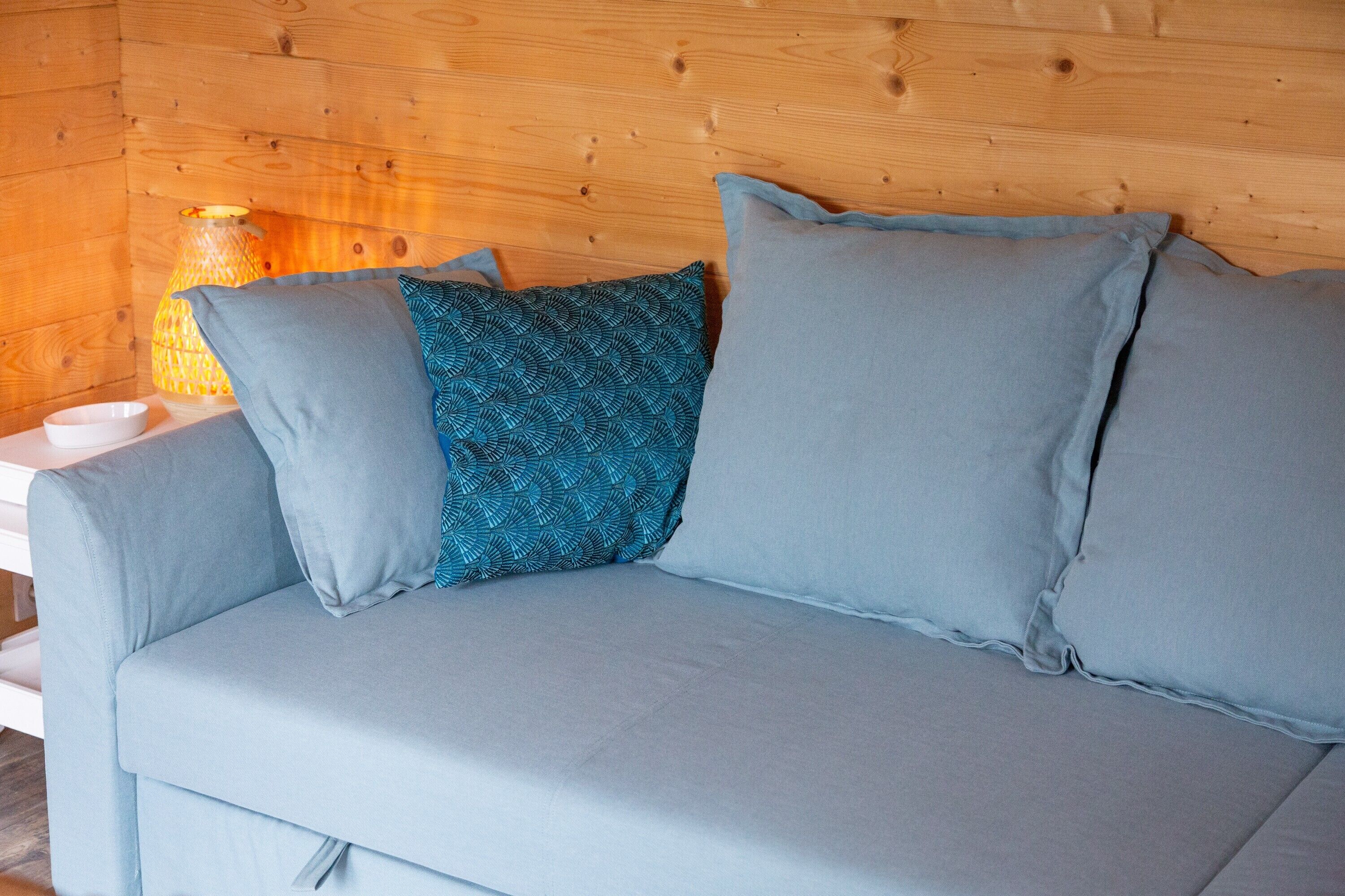 Un sofá azul claro con almohadones estampados y lisos, un cuenco blanco y una pared de madera con una lámpara.